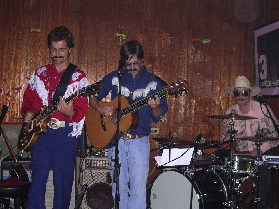 Three-piece band on stage in western wear — two guitars and a drummer in a cowboy hat.