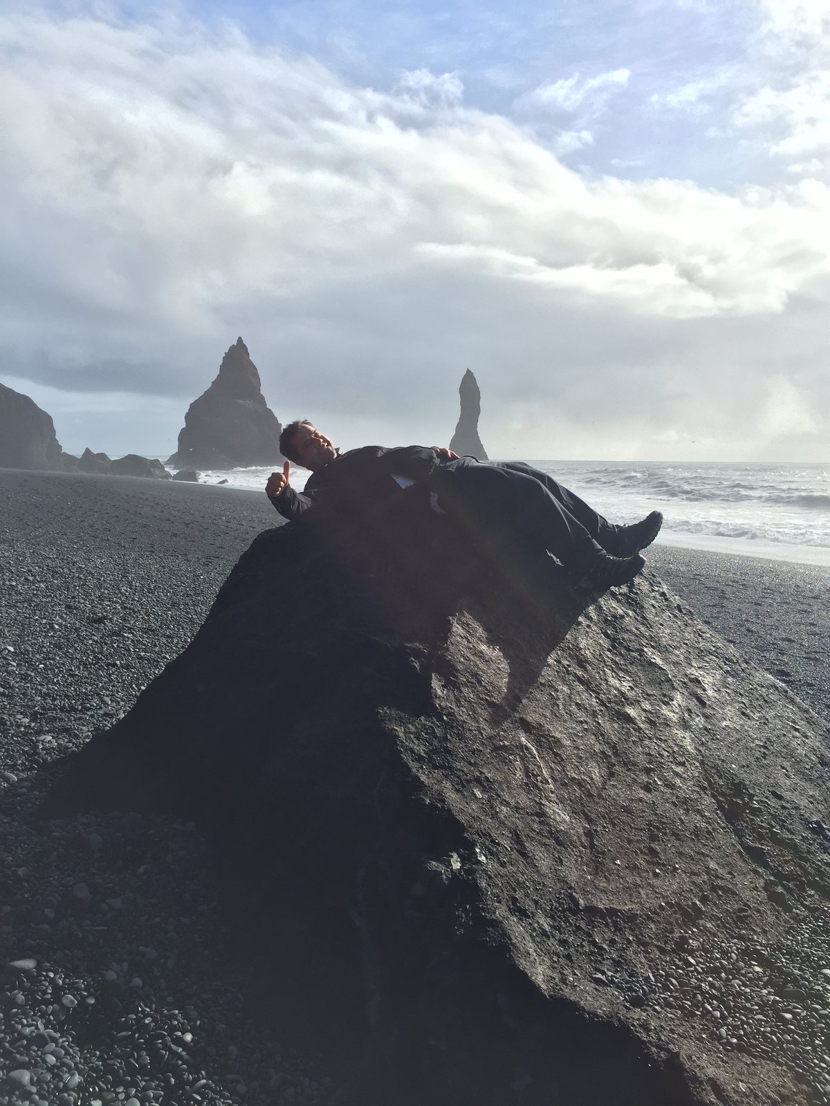Lying flat on a black boulder on a black sand beach in Iceland, giving a thumbs up, sea stacks behind.