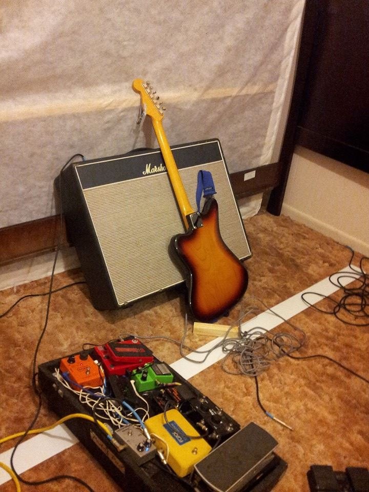 Sunburst Jazzmaster leaning against a Marshall combo amp, pedalboard and cables sprawled on the floor.
