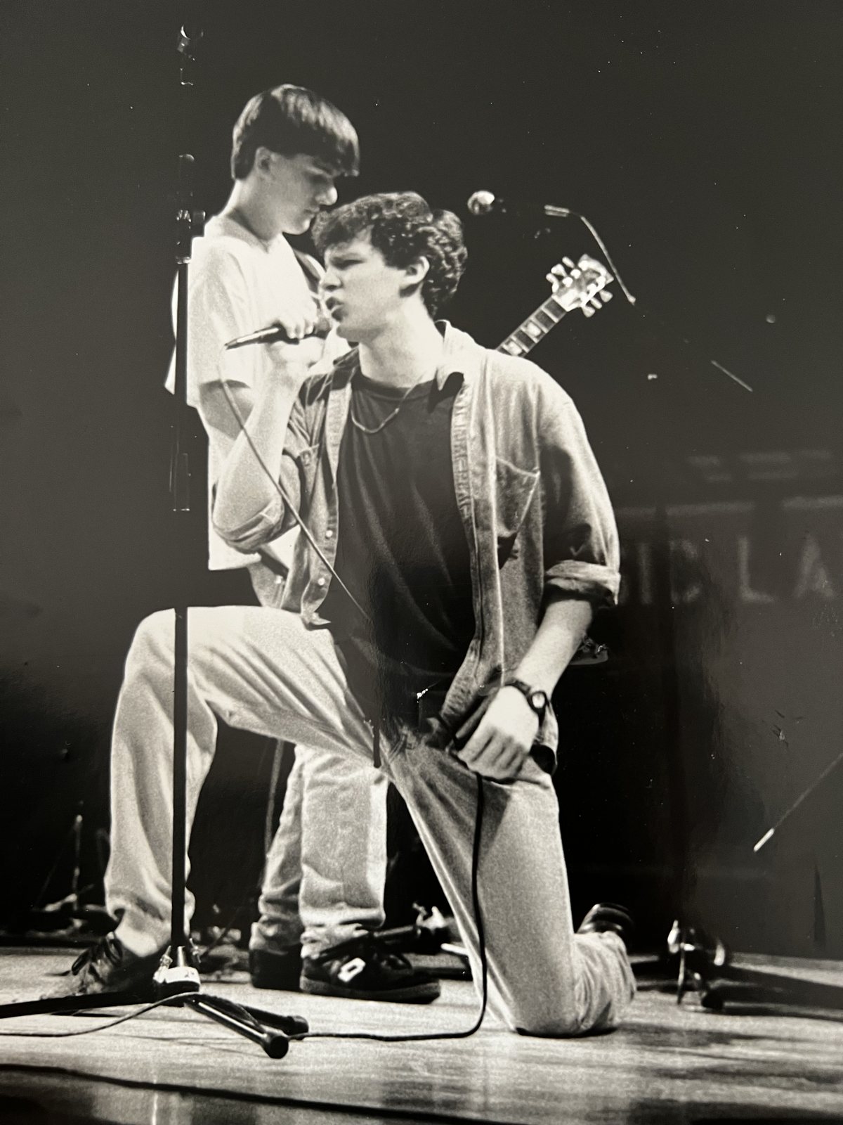 Black-and-white photo of two teenagers performing on stage, one down on a knee singing into a microphone.