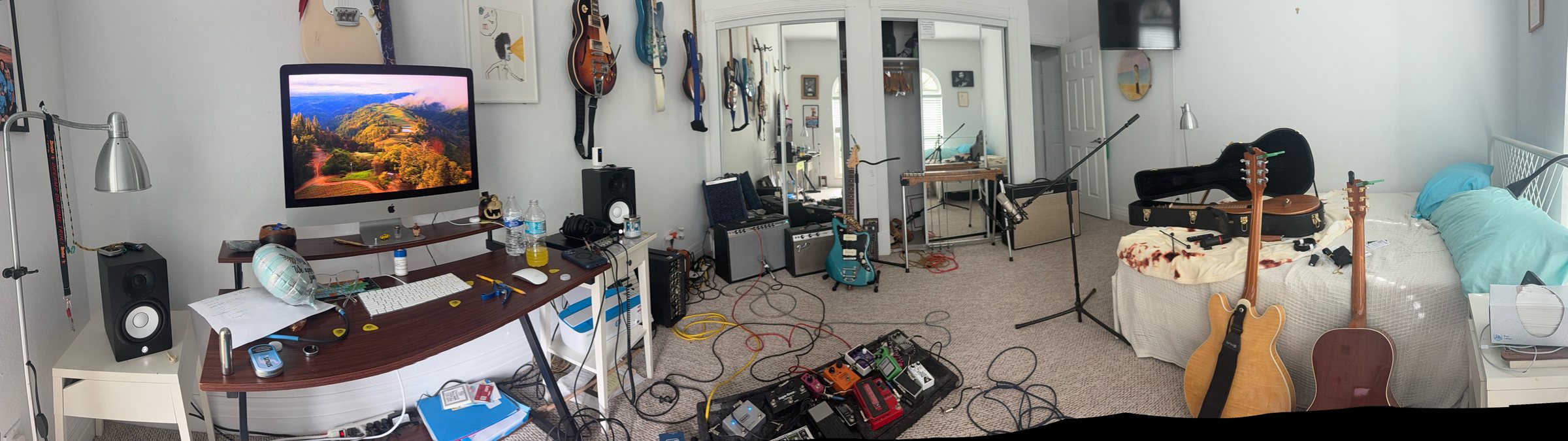 Panorama of the home studio: guitars on the walls, amps, a pedalboard mid-tangle, and a bed covered in gear.